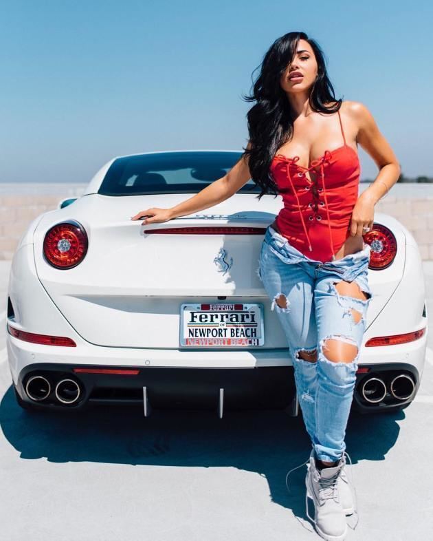 Ana Cheri looking fierce while holding a car's trunk and wearing a red swimsuit, ripped jeans, and white sneakers