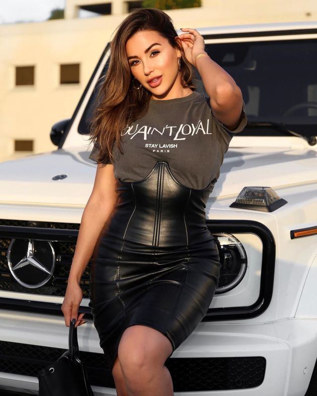 Ana Cheri leaning in a white car and smiling with her hand touching her hair while wearing a black dress, round earrings, and carrying a bag