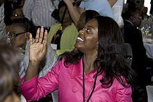 Ana Paula dos Santos smiling while looking at something, waving her hand, and wearing a pink blouse