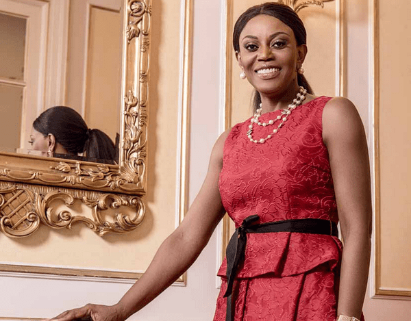 Ana Paula dos Santos smiling while wearing a red sleeveless dress with a black ribbon belt, pearl necklace, and pearl earrings