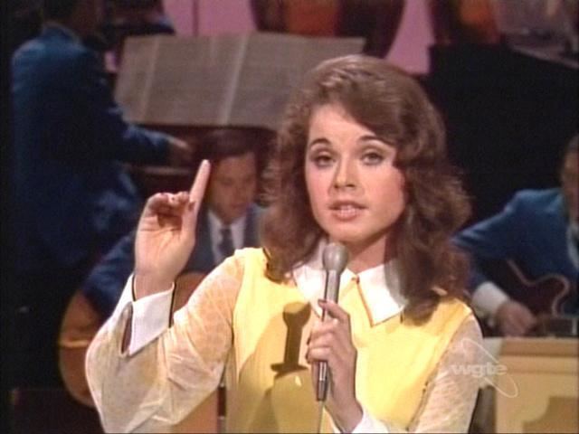 Anacani holding the microphone while pointing her finger upward and wearing a yellow blouse
