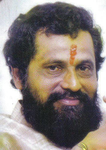 Anand Dighe with a tight-lipped smile, mustache, and beard while wearing a white and beige checkered polo