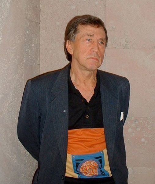 Anatoli Bugorski with a serious face while his hands are at his back and wearing a black coat over a multi-colored polo shirt.