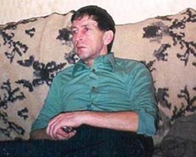 Anatoli Bugorski with a serious face while lying on the couch and holding his own hands, wearing green folded long sleeves and black pants.