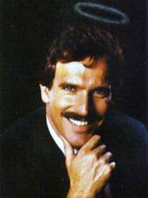 Andrew Clarke is smiling, has black hair and a mustache, a white circle on left above his head, his right hand on his chin, wearing white long sleeves under a black suit.