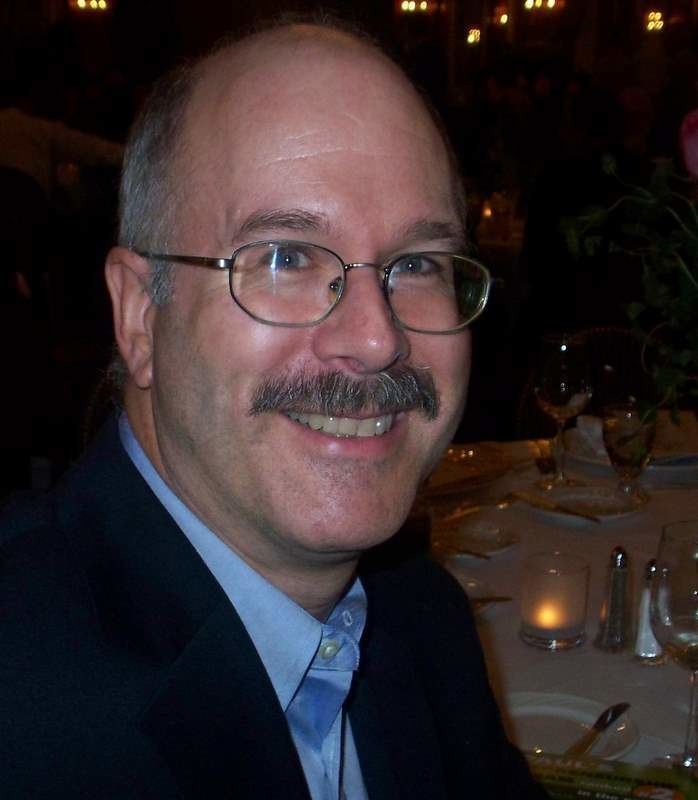 Andrew Clarke is smiling, has white hair, a shaved beard, and a mustache, sitting down beside a white table with plates, a glass, a flower, a candle inside a glass, a bottle of salt and pepper, and a knife,  he is wearing eyeglasses, blue long sleeves under a black suit.