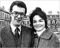 Andrew Russell, 15th Duke of Bedford, smiling and wearing eyeglasses, long sleeves, and coat while his wife wearing a coat and turtle-neck blouse