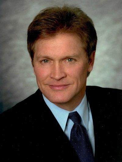 Andrew Stevens with a tight-lipped smile while wearing a black coat, blue long sleeves, and dark blue necktie