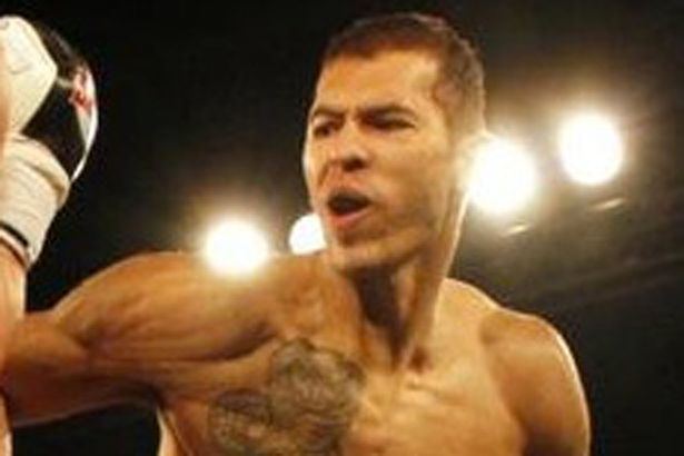 In a dark stadium with a large spotlight, Andre Tate is serious, mouth half open, fighting left arm throwing a punch, has brown hair, with a snake tattoo on his right chest, top naked.