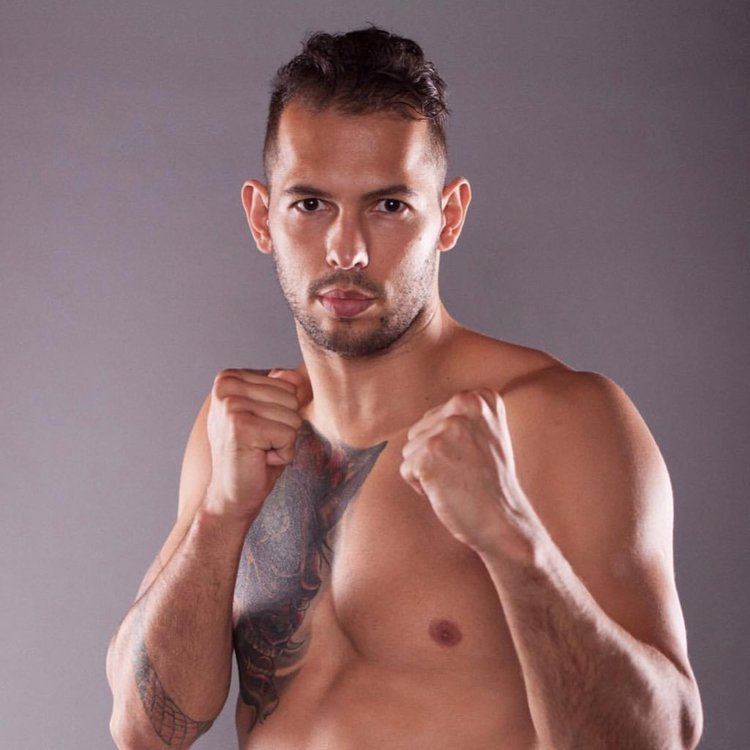 In a gray background, Andrew tate is serious, with his fierce look, standing, fighting pose, both hands up fist closed, has tattoo on his chest and right arm, black hair mustache and beard top naked.