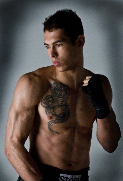 In a gray backdrop, Andrew Tate is serious, standing posing with his right arm down, and left hand up closed, looking at his right, has black hair, a snake tattoo on his right chest, wearing a black gloves and black shorts.