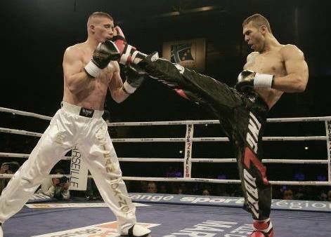 In a boxing ring, from left, a man is serious, standing, guarding a kick, fighting, both legs apart, has brown hair, wearing a black gloves wrapped with boxing tape and a white pants with white shoes,at the right, Andrew Tate is serious, fighting in a ring, throwing a straight kick with his right foot on the face of the man, left foot on the ground, left hand bent over his stomach, has black hair, wearing a black gloves wrapped with white boxing tape a black pants and red shoes.