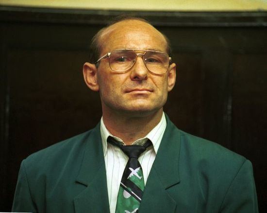 Andrzej Kolikowski wearing a green coat, white striped long sleeves, black and green necktie, and eyeglasses