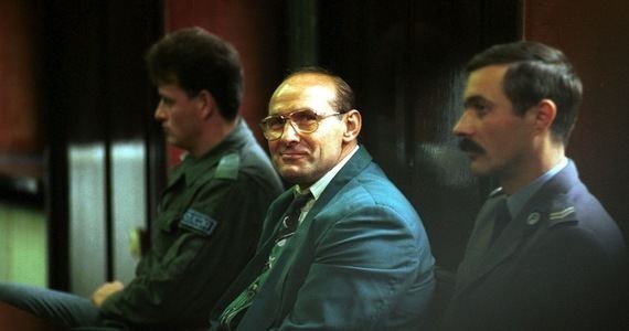 Andrzej Kolikowski smiling and sitting on the chair with two men beside him while he is wearing a green coat, long sleeves, necktie, and eyeglasses