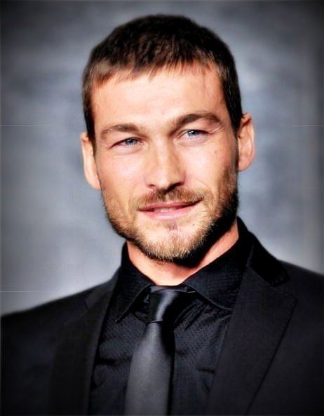Andy Whitfield smiling, with beard and mustache, and wearing a black suit, black long sleeves, and black tie.