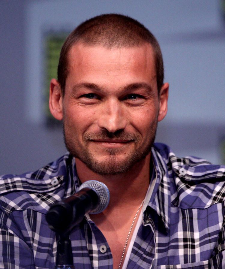 Andy Whitfield smiling with a microphone in front of him, with beard and mustache, wearing a necklace and a blue checked polo shirt.
