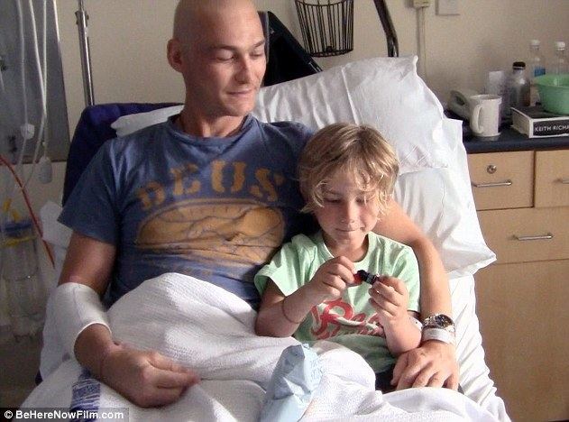 Andy Whitfield smiling while lying on a hospital bed with his son Jesse, with a bald head, and wearing a blue shirt.