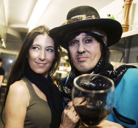 Angela Nicoletti wearing a black scarf and green top with a man wearing a black hat, a blue shirt, and holding a wine glass.