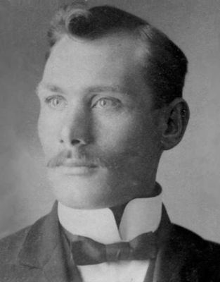 Angelo Hays with a serious face while looking at something, with a mustache, wearing a black coat over white long sleeves, and a black bowtie.