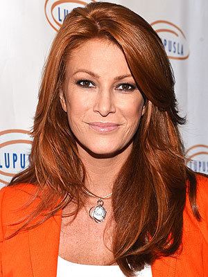 Angie Everhart smiling with orange-brown hair and wearing a necklace and white blouse under a bright red coat