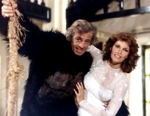 Jean-Paul Belmondo and Raquel Welch are smiling and swinging on a woody vine while Jean-Paul is wearing a gorilla costume and Raquel Welch is wearing a white long sleeve dress in a scene from the 1977 film, L'Animal