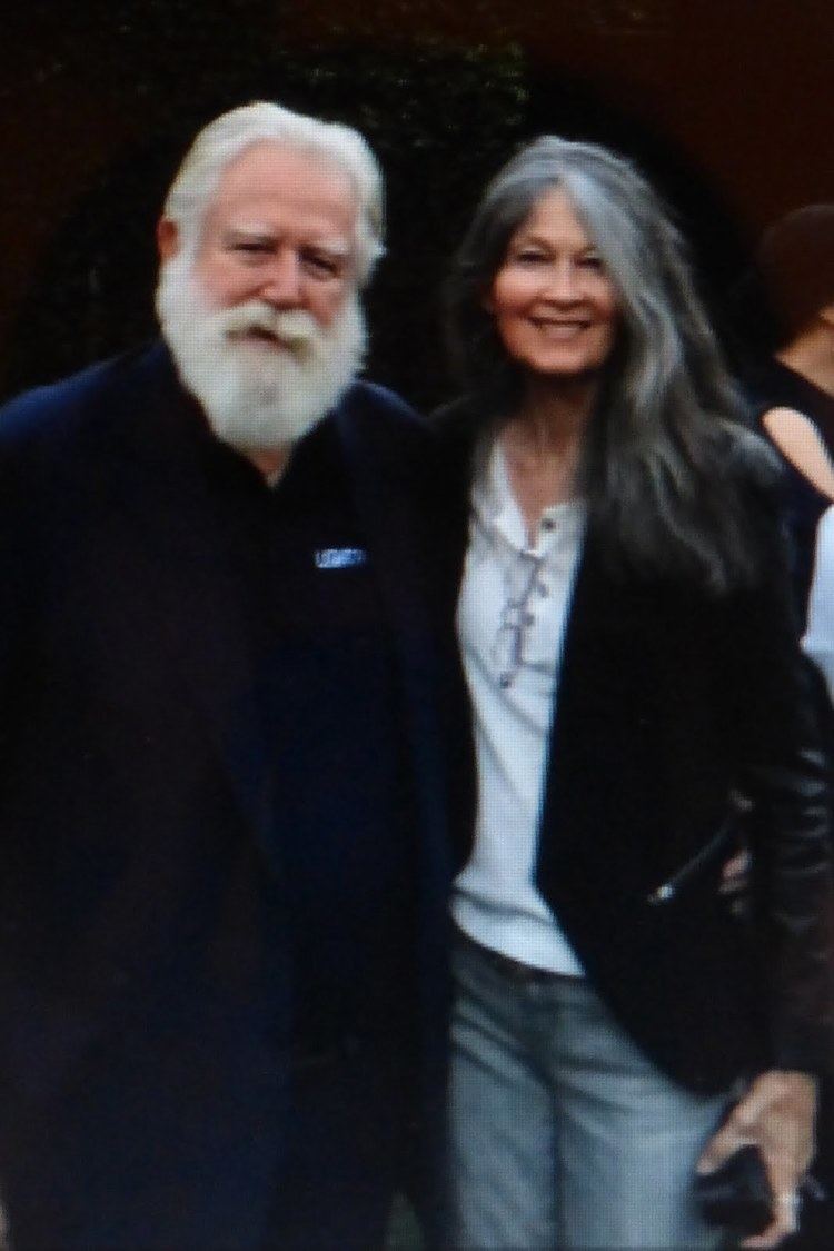 Anitra Ford smiling and wearing a black coat, white t-shirt, and pants while the man beside her with mustache and beard wearing a black suit