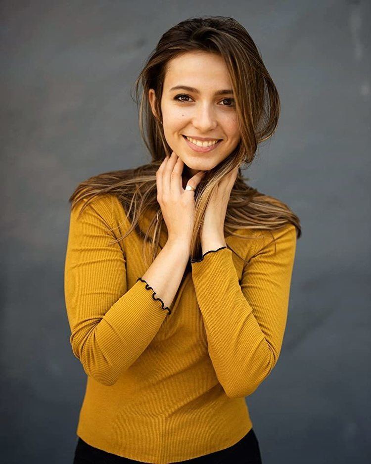 Anjelica Bette Fellini smiling while hands on her neck and wearing a mustard yellow long sleeve blouse and ring