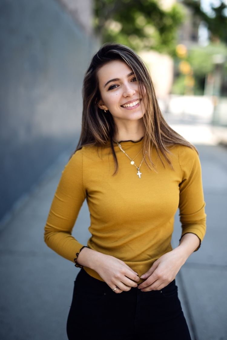 Anjelica Bette Fellini smiling while wearing a mustard yellow long sleeve blouse and necklace with cross pendant
