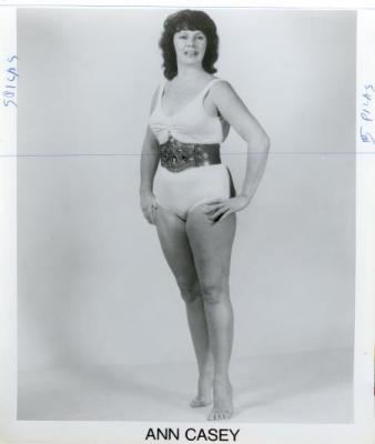 Ann Casey posing with her hand on her waist and wearing a white wrestling attire with her title belt.