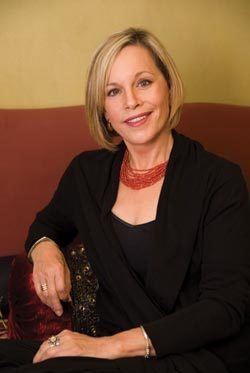 Ann Maurice smiling, with short blonde hair and wearing a red necklace and black suit.