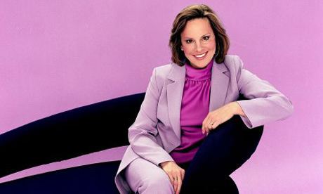 Ann Maurice smiling, with short wavy hair, wearing a purple shirt and a blazer while sitting on a chair.