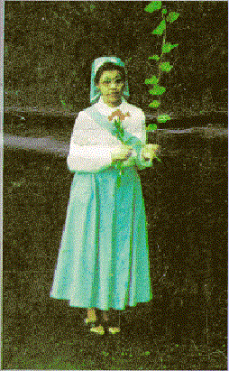 Anna Ali holding leaves of some plant and wearing a mint green veil, white long sleeve blouse, mint green skirt, and black sandals.