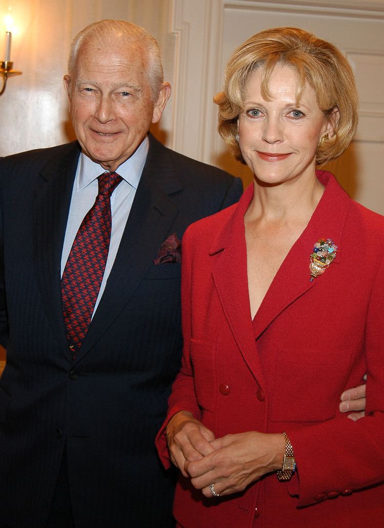 In a white room with electric wall candle lamp and a white door, from left, Rupert Murdoch is smiling, standing, with his hands at the back waist of Anna, has bald top and white side hair, wearing a light blue polo with black and red printed necktie under a black coat with a maroon scarf on left chest pocket, at the right, Anna Murdoch Mann is smiling, standing, with her hands together in her stomach, has short blond hair wearing a silver ring gold watch, and a red long coat with large brooch on its left chest.