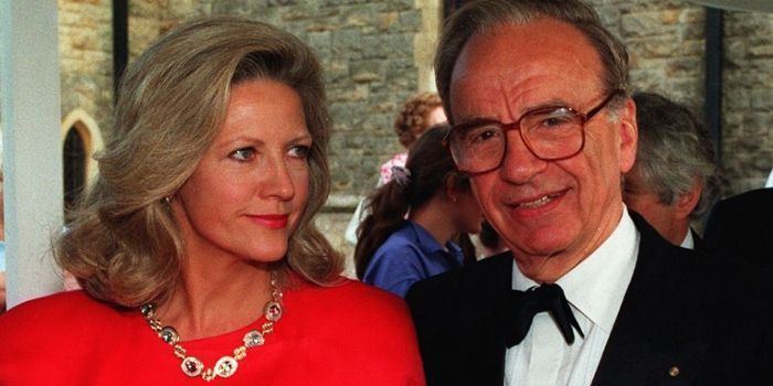 In front of large bricks wall with people at the back, from left, Anna Murdoch Mann is smiling, standing, looking to Rupert, has brown blonde wavy hair, wearing red lipstick a gold necklace and a red dress, at the right Rupert Murdoch is smiling, standing has dark brown hair wearing eyeglasses a white polo with black bowtie under a black coat.
