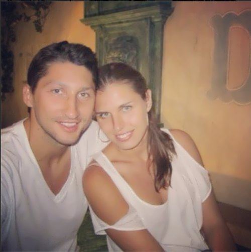 Anna Petrakova smiling with Nikita Kurbanov while leaning on him. Anna is wearing a visible cleavage white shoulder cut-out blouse, and silver round earrings while Nikita is wearing a visible cleavage white shirt