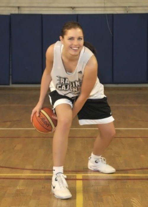 Anna Petrakova smiling while dribbling a ball, having her hair tied with a blue foam mattress in the background, wearing a white jersey with the printed word "RAGIN CAJUNS", jersey number 13, and Adidas logo, paired with black and white shorts, and white socks under white shoes