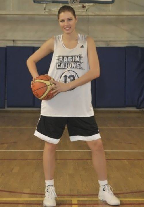 Anna Petrakova smiling while holding a ball with her two hands, having her hair tied with a basketball ring and blue foam mattress in the background, wearing a white jersey with the printed word "RAGIN CAJUNS", jersey number 13, and Adidas logo, paired with black and white shorts, and white socks under white shoes