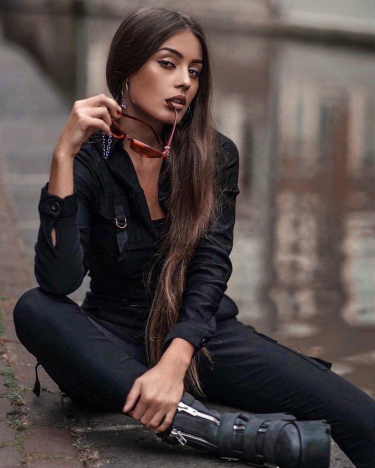 Anna Zapala sitting on the ground while holding red sunglasses and wearing a black long sleeve blouse, black pants, and black shoes