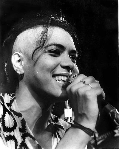 Annabella Lwin singing, holding a mike on her left hand, two rings a black thread tied, faded hairstyle, wearing black and white top