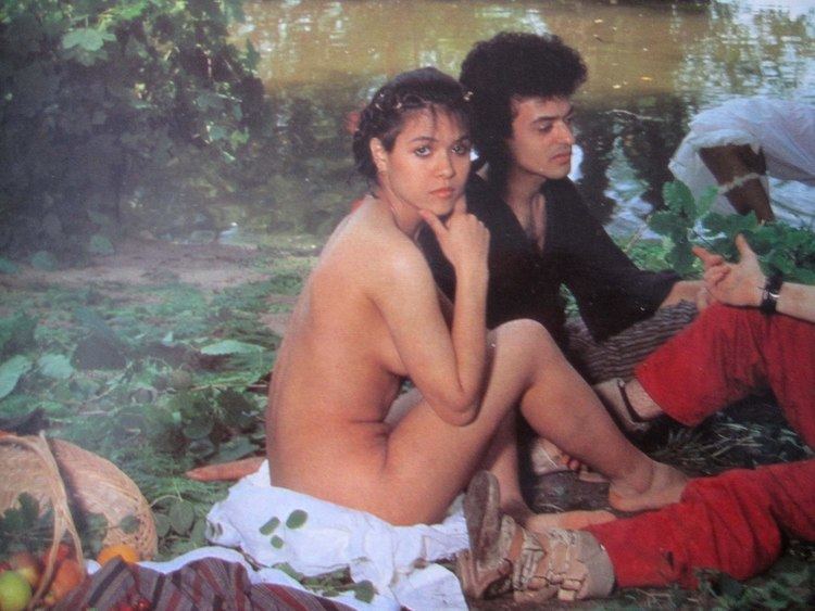 Annabella Lwin seating in ground with her friend. Annabella twin is looking serious , taking support of her face with her hand on chin, blonde hairs and naked