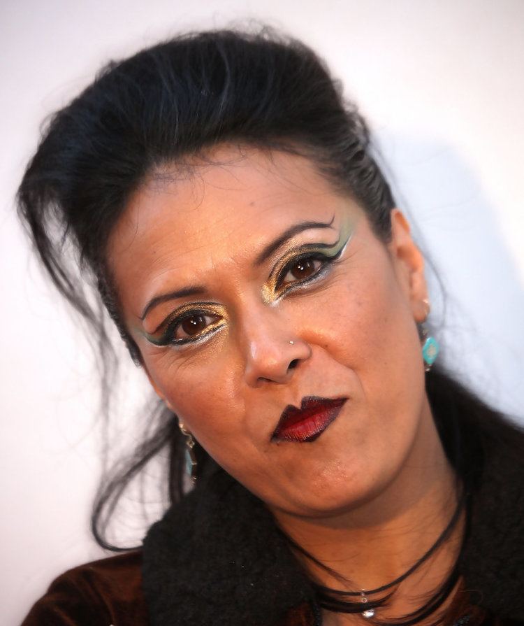 Annabella Lwin with a tight lipped smile while wearing blue hearings, small nose ring and black necklace with blonde hairs
