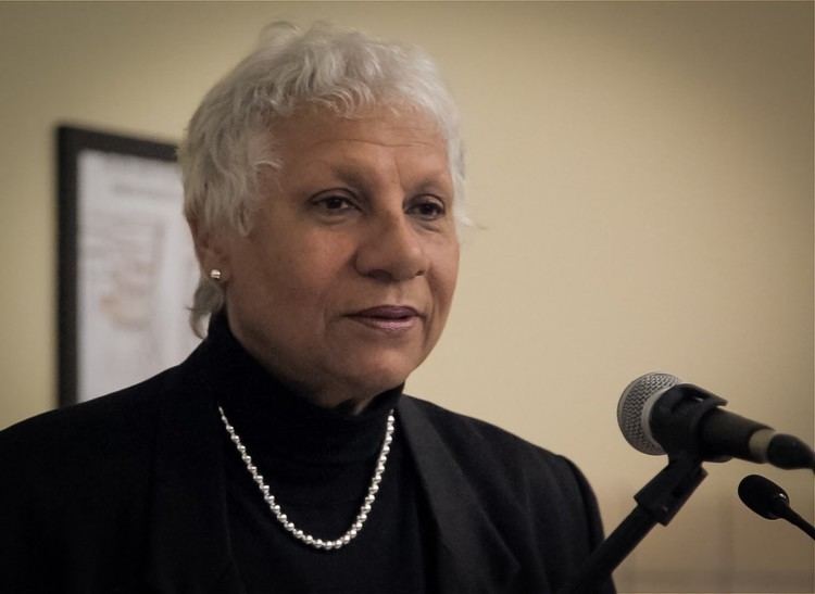 Anne Cools looking at something with a microphone in front of her. Anne with white short wavy hair is wearing earrings, a necklace, and a black turtle neck blouse under a black coat