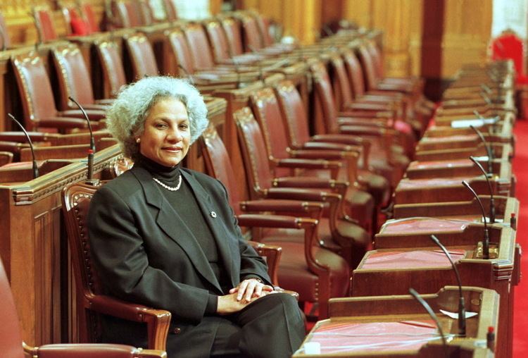 Anne Cools smiling while sitting on the tribune chair and her hands together, with white short curly hair, and wearing a necklace, earrings, black pants, and a black blouse under a black coat