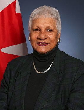 Anne Cools with a tight-lipped smile and white short hair, behind her is the Flag of Canada, and she is wearing earrings, a necklace, and a black turtle neck blouse under a black coat
