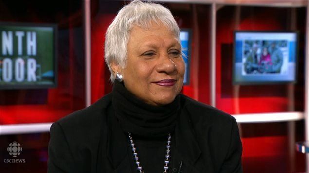 Anne Cools with a tight-lipped smile and white short hair while wearing earrings, a necklace, and a black turtle neck blouse under a black coat