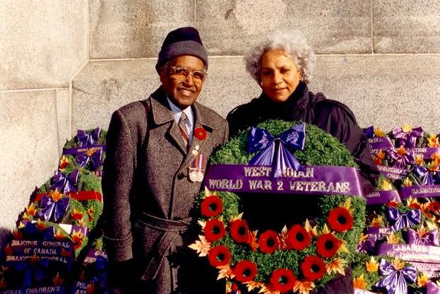 A man and Anne Cools are smiling while posing in front of a standing bouquet for the WWII veterans. The man standing beside Ann is wearing a gray beanie, eyeglasses, and blue long sleeves under a gray and red striped necktie and a gray trench coat with medals on it, while Anne is wearing a black coat