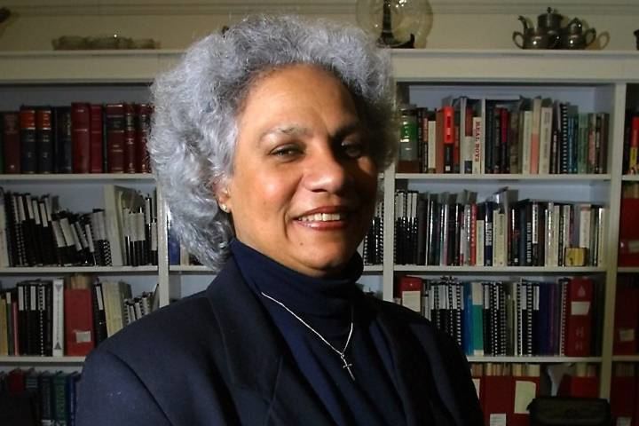 Anne Cools smiling with white short curly hair and bookshelves behind her while wearing earrings, necklace, and black turtle-neck blouse under a black coat