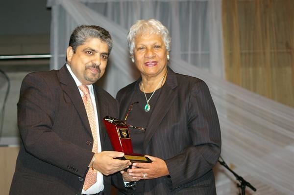 Anne Cools and the man beside her are smiling while holding a trophy. Anne with white short wavy hair is wearing earrings, a necklace, a ring, and a black blouse under a black coat while the man beside her with a mustache and beard is wearing a white long sleeve under a peach necktie, and a black coat