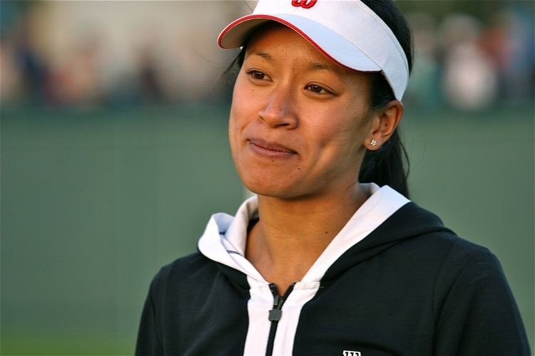 Anne Keothavong with a tight-lipped smile while looking at something and wearing a black and white jacket and white and red cap