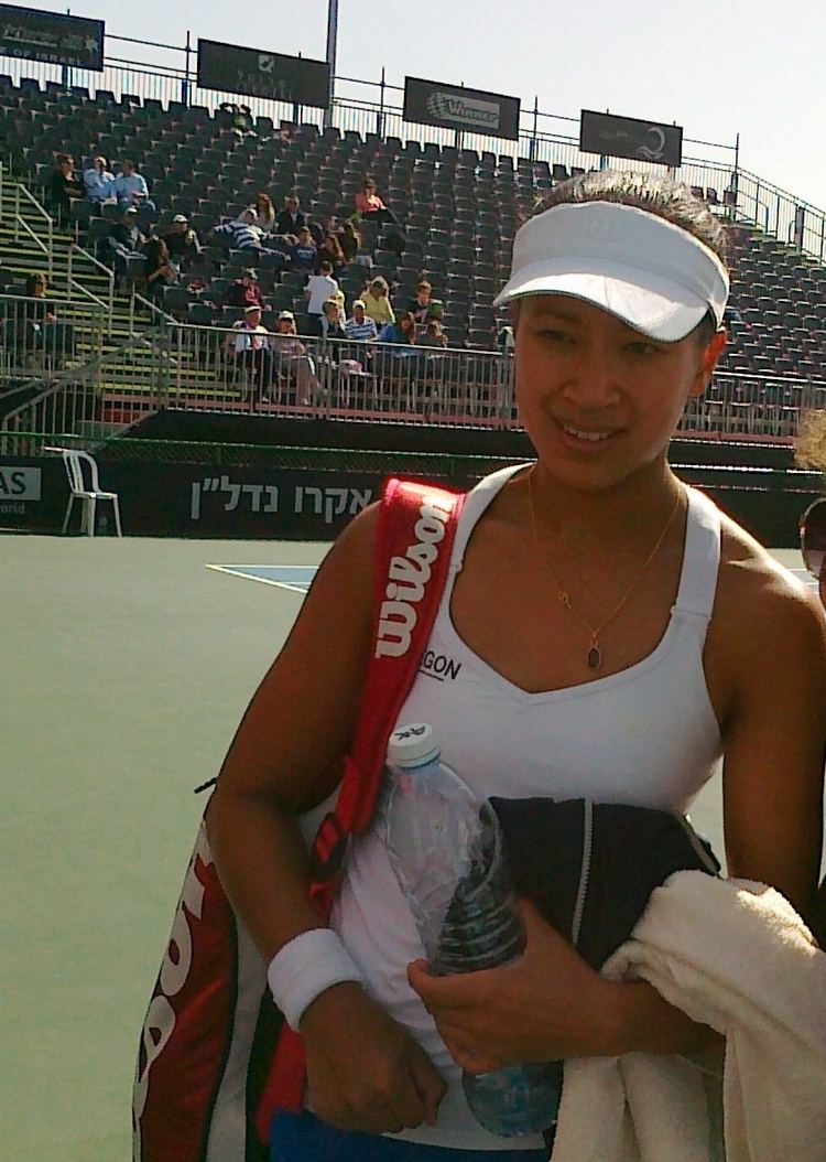 Anne Keothavong smiling and carrying a red bag, water bottle, and towel at the Fed Cup in Eilat, Israel while wearing a white tank top